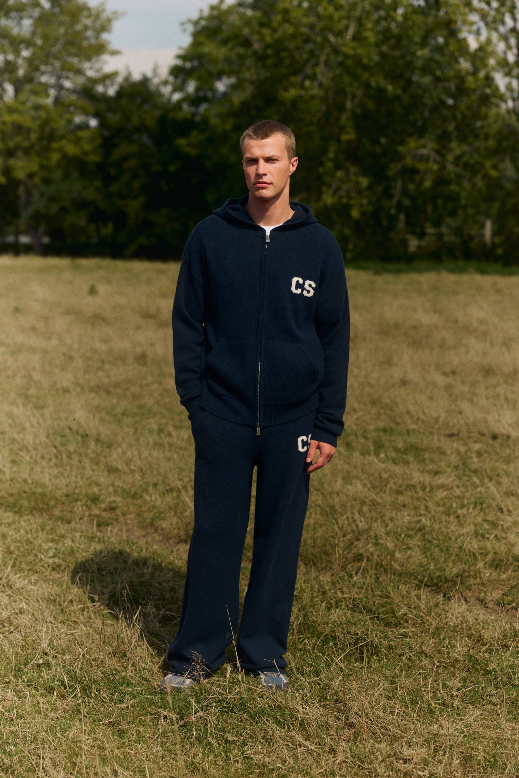 Man wearing a navy CASPARA woolknit zip-up hoodie and straight-leg pants in a grassy field.